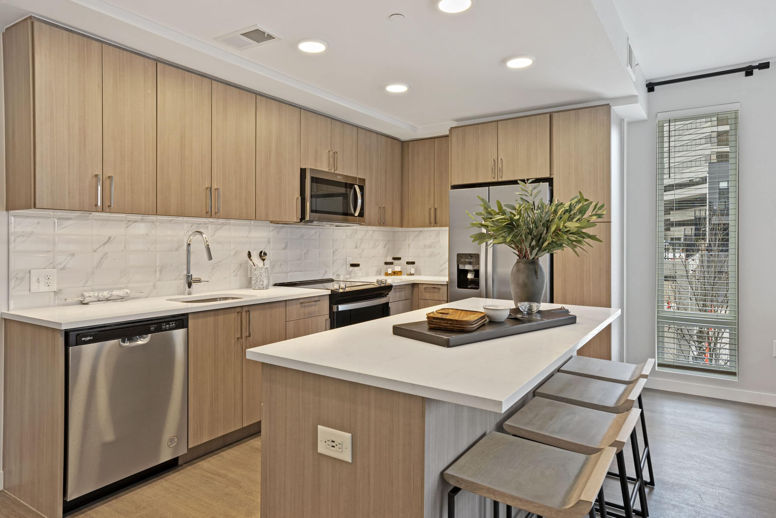 Modern kitchen with light wood cabinets, white countertops, stainless steel appliances, and a central island with bar stools. A vase with green branches and a tray decorates the island. Large window provides natural light.