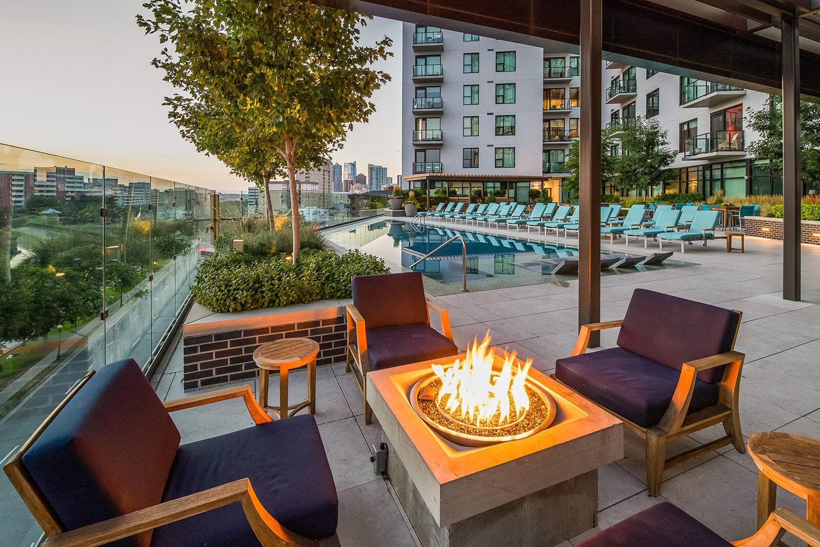 Outdoor seating with navy cushioned chairs surrounds a fire pit on a modern patio at 1000 Speer, overlooking a pool with lounge chairs. Apartment buildings and a city skyline are visible in the background at sunset.