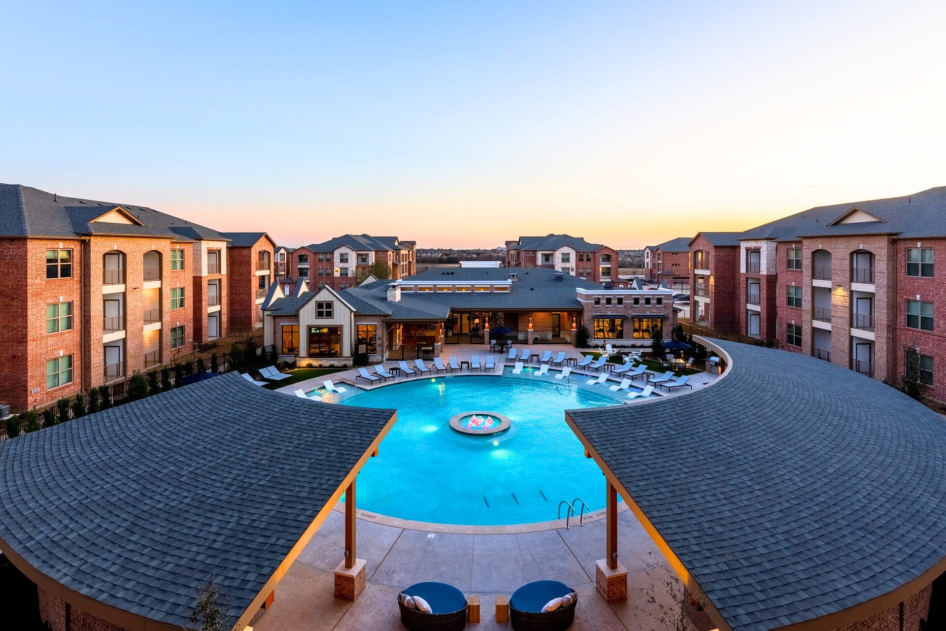 A large, circular outdoor swimming pool at Windsor Castle Hills is surrounded by lounge chairs and modern apartment buildings at sunset, with a clear sky and warm lighting.