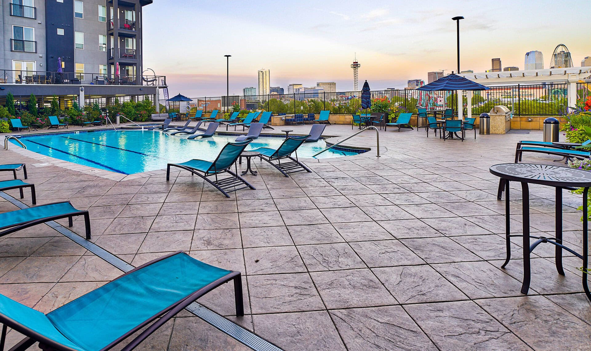 Outdoor pool area with blue lounge chairs and tables on a tiled deck, surrounded by a fence. The city skyline glows at sunset, with tall buildings and the clear sky reflecting the sleek style found at Element 47.