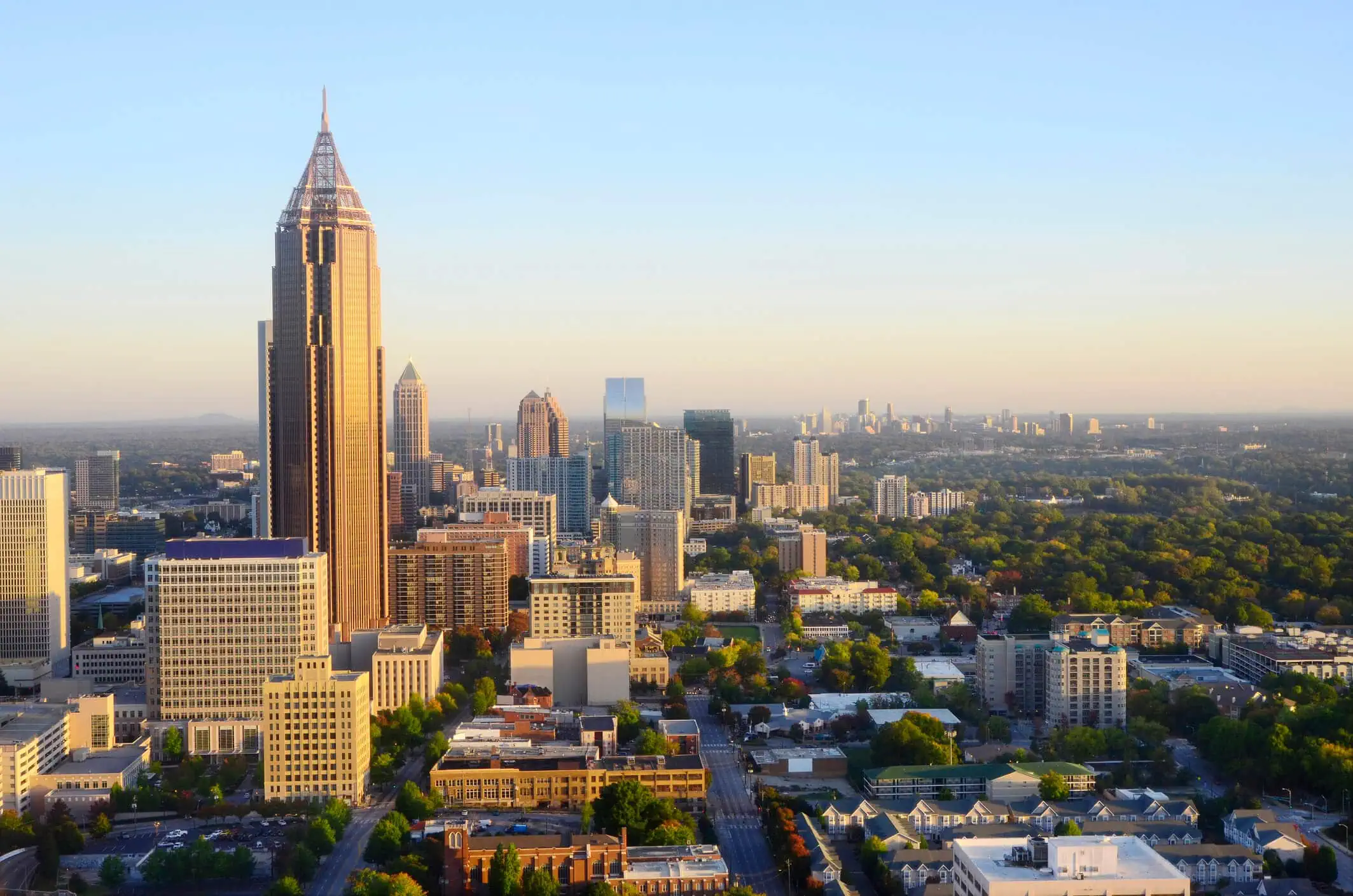 Aerial view of downtown Atlanta, Georgia, featuring tall skyscrapers, green trees, and buildings under a clear blue sky in the early morning light.