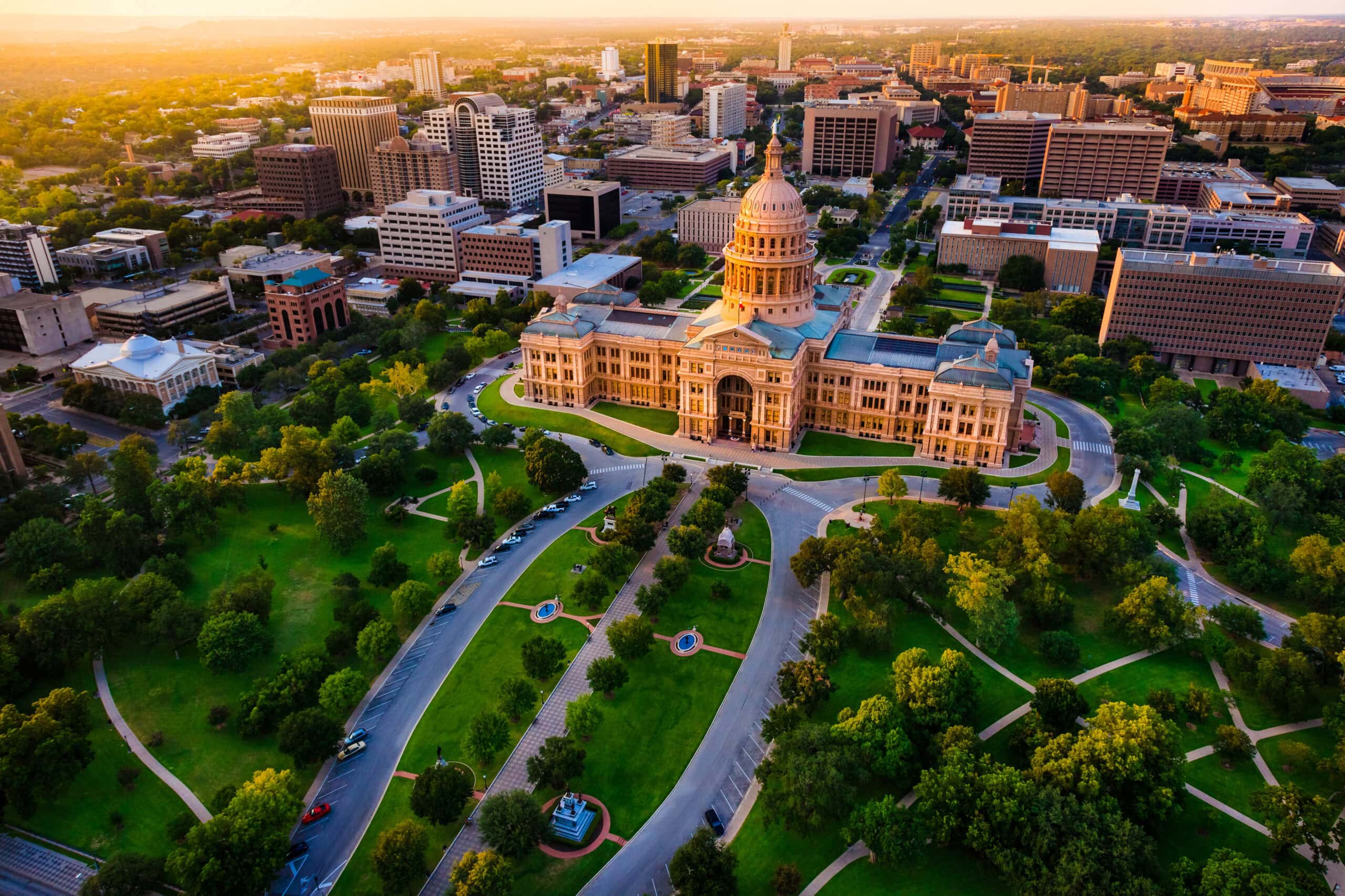 Aerial view of the Texas State Capitol building in Austin, surrounded by green trees and gardens, with the city skyline and buildings in the background at sunset.
