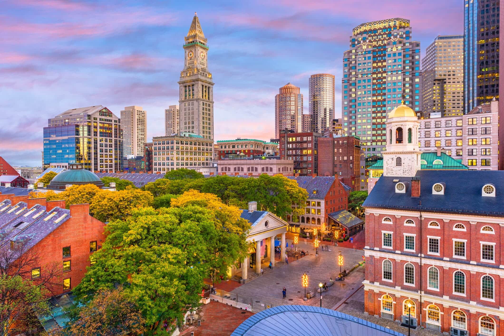 Boston cityscape at sunset, featuring historic Faneuil Hall in the foreground, leafy trees, and modern skyscrapers like The Victor rising in the background under a colorful pink and blue sky.