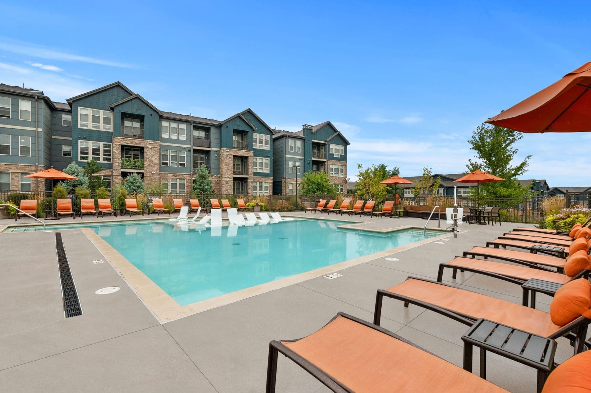 View of Windsor at Pinehurst, a modern apartment complex featuring a large outdoor swimming pool surrounded by lounge chairs and orange umbrellas under a bright blue sky. Some lounge chairs are set partially in the water for ultimate relaxation.
