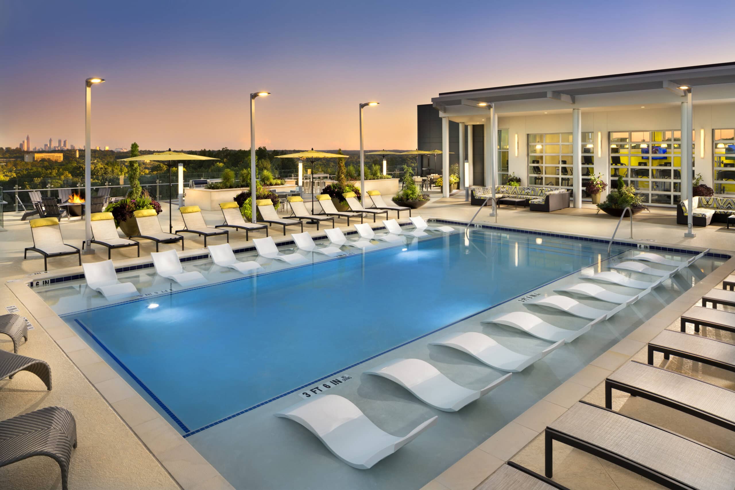 Modern rooftop pool at sunset at Windsor Encore, with white lounge chairs partially submerged in the shallow end, surrounded by sun loungers, potted plants, and a seating area. City skyline visible in the distance.