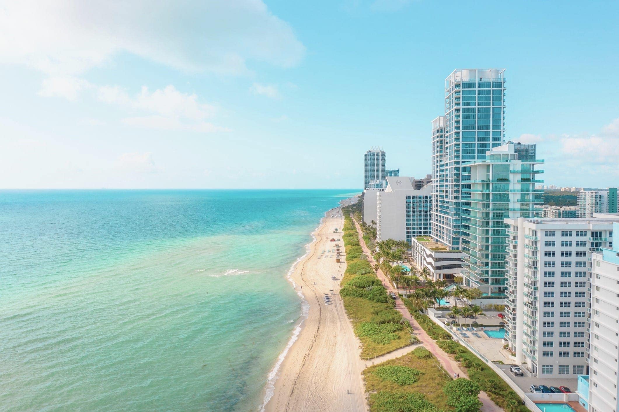 High-rise buildings line a sandy beach with turquoise ocean water on a sunny day. The coastline curves into the distance under a blue sky with scattered clouds.