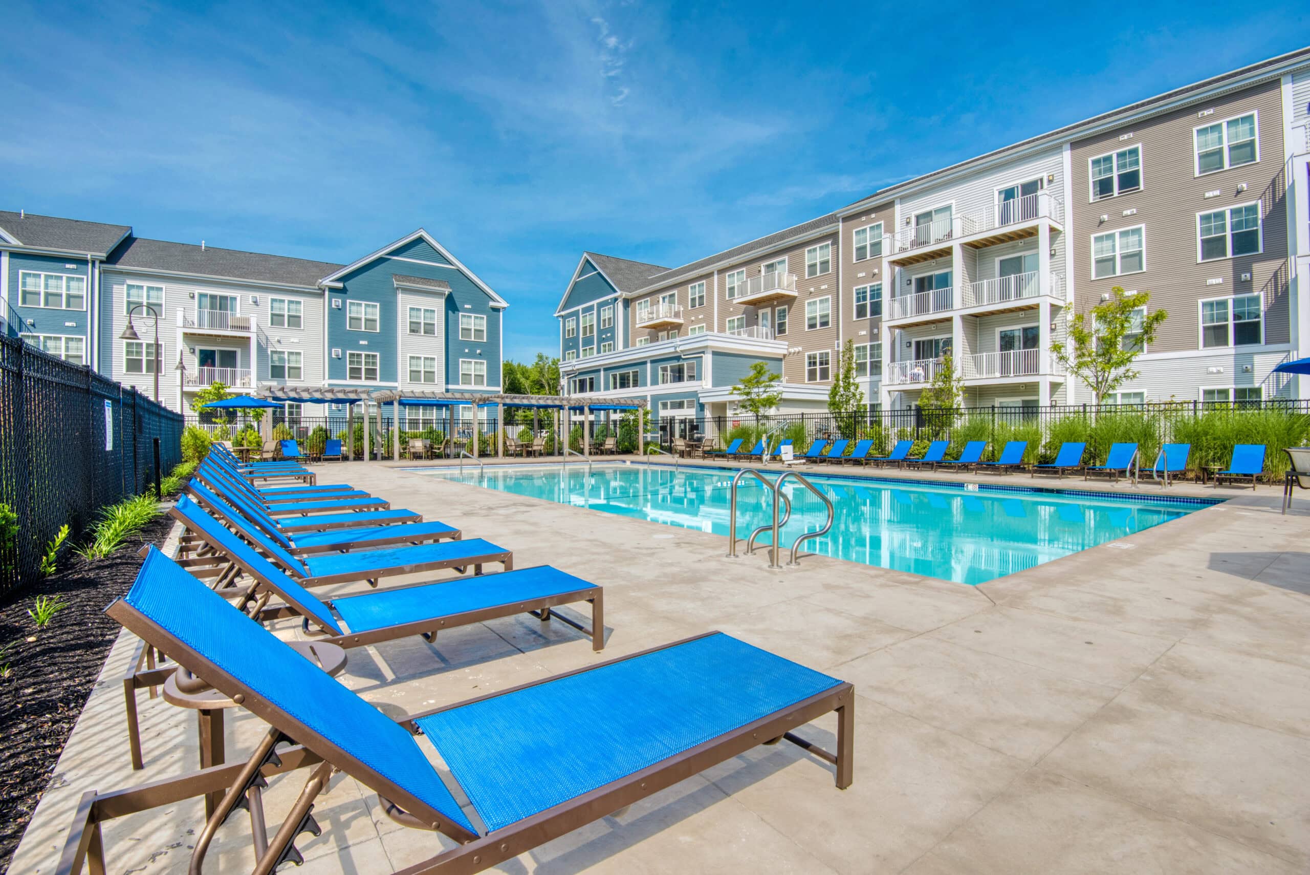Outdoor swimming pool surrounded by blue lounge chairs at Windsor at Hopkinton, with modern apartment buildings in the background under a clear blue sky.