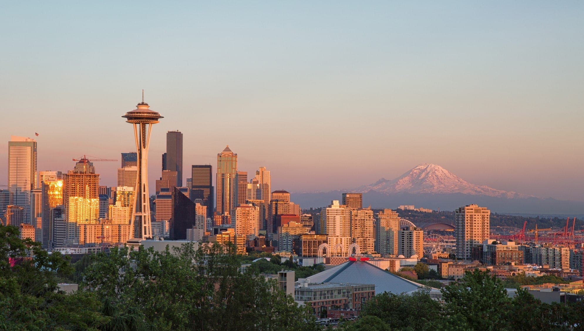 Seattle skyline at sunset with the Space Needle and downtown skyscrapers in view; Mount Rainier is visible in the background under a clear sky. Trees and city buildings are in the foreground.