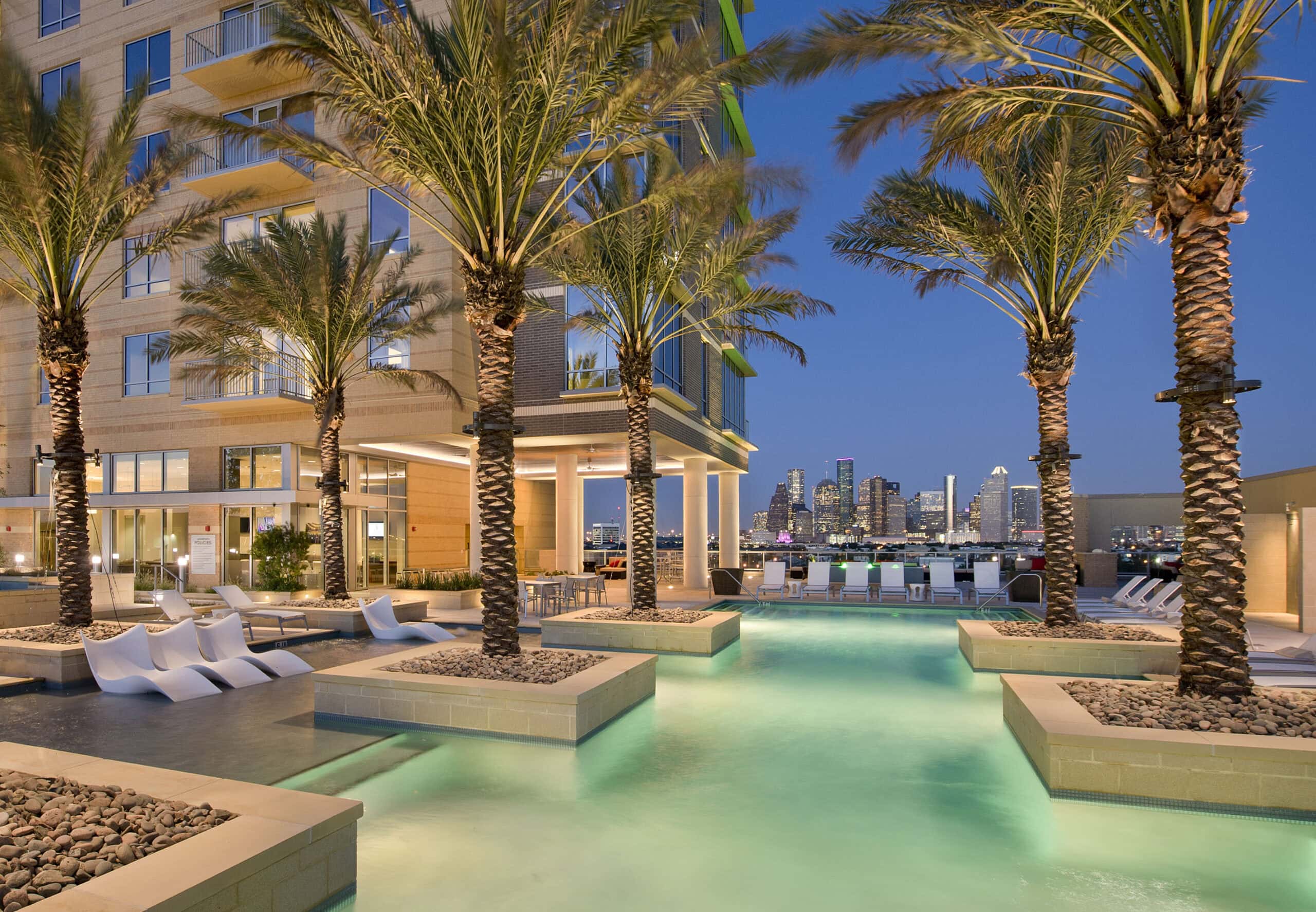 A luxurious rooftop pool at The Sovereign at Regent Square features palm trees, lounge chairs in the water, and a stunning view of the modern city skyline at dusk, with building lights reflecting off the shimmering surface.