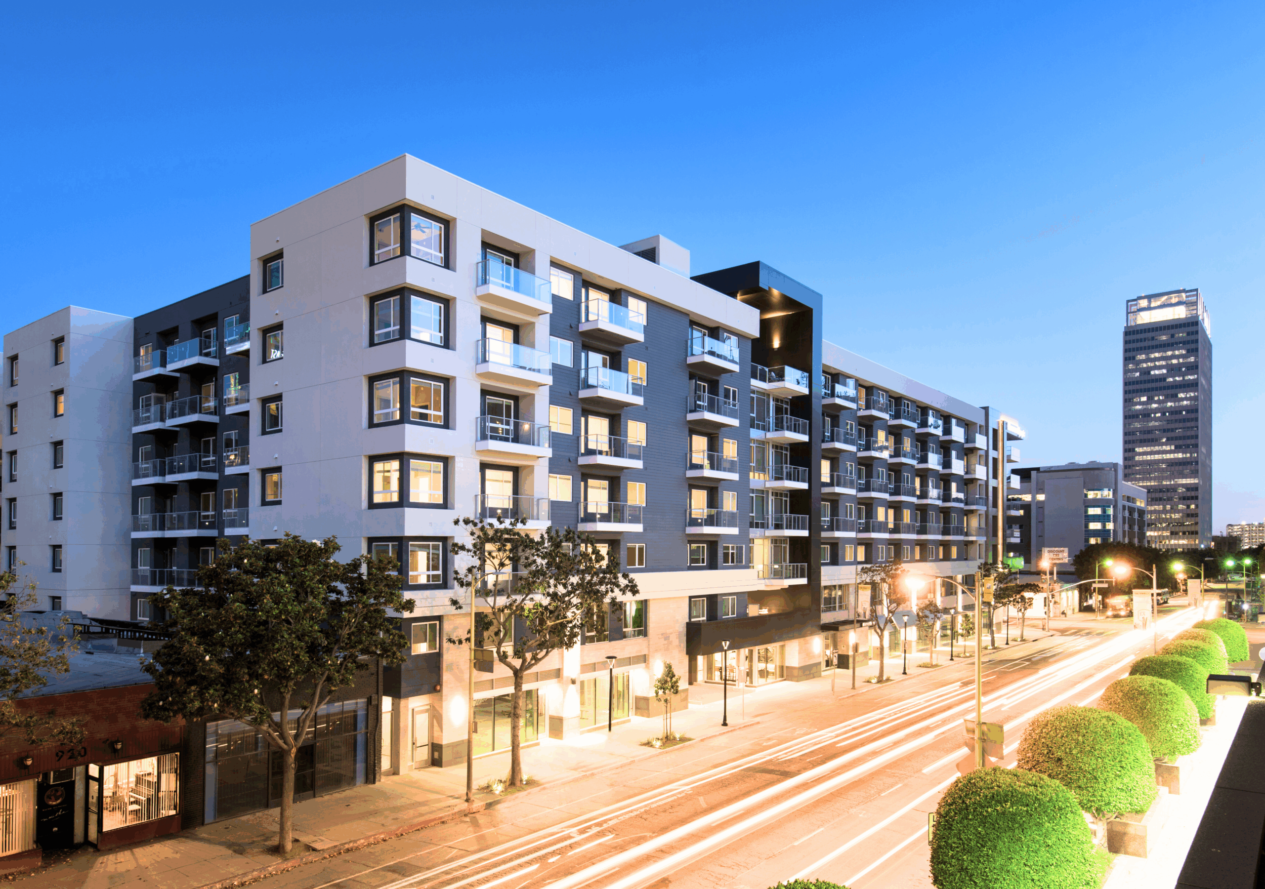 Modern mid-rise apartment building, Olympic by Windsor, features illuminated windows at dusk, surrounded by trees and streetlights. Light trails from passing cars enhance the scene, with a tall office building in the background against a clear sky.