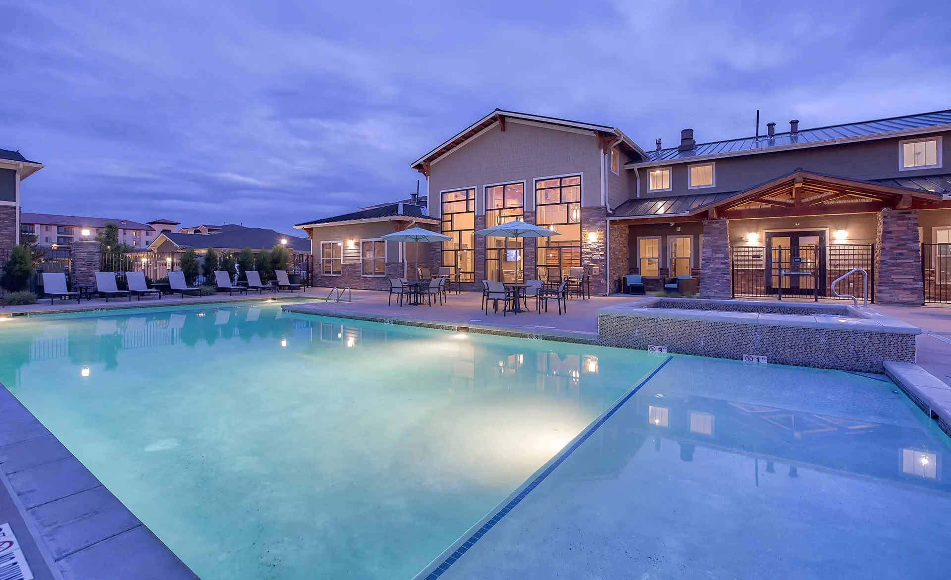 A large outdoor swimming pool with lounge chairs and tables with umbrellas sits in front of the modern two-story Retreat at the Flat Irons building at dusk, as warm lights illuminate the windows and patio under a cloudy sky.