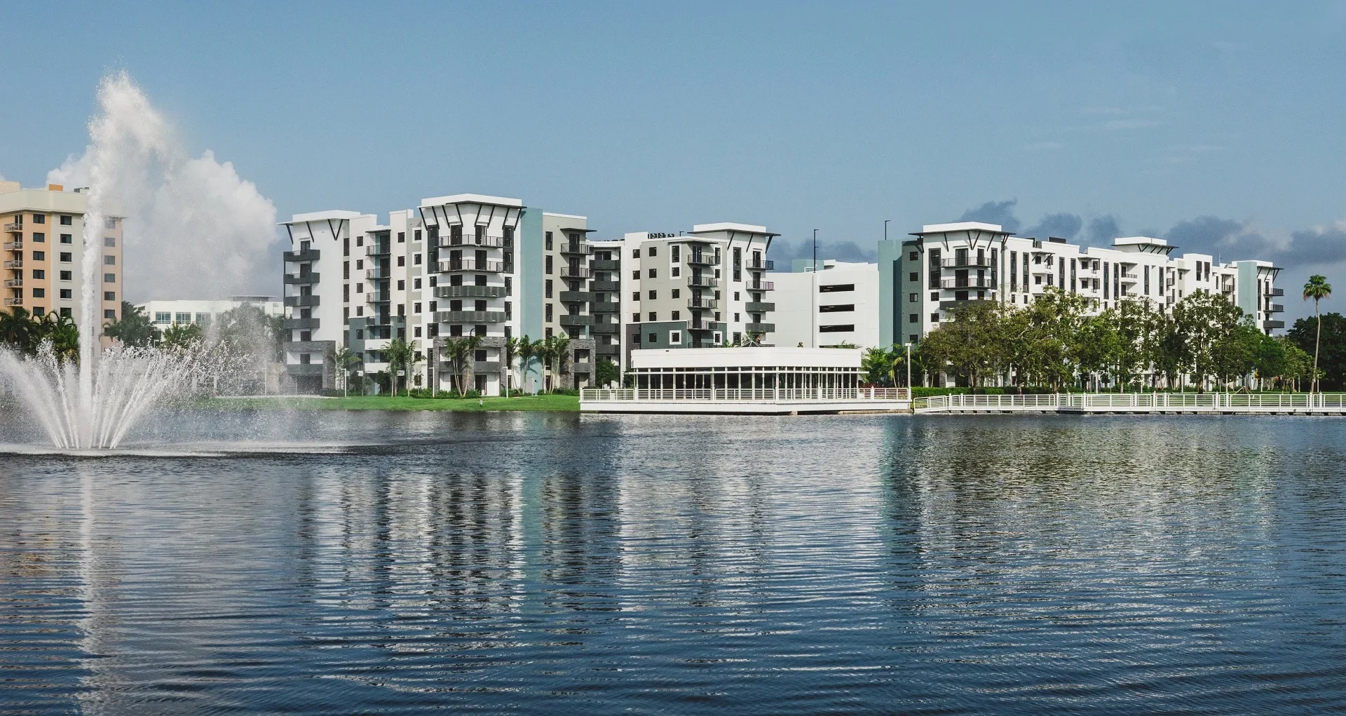 Modern mid-rise apartment buildings with white and gray exteriors, including Windsor Cornerstone, are reflected in a calm lake. A large water fountain sprays on the left, framed by a blue sky and trees along the waterfront.